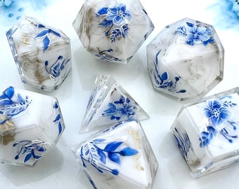May include: A collection of seven dice with a floral design. The dice are clear with a white interior and blue floral accents. Some dice have gold leaf details. The dice are in various geometric shapes, including cubes, pyramids, and multi-sided shapes.