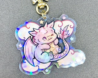 May include: A holographic dragon keychain with a gold star clasp. The dragon is pink and purple, holding a gem, and surrounded by clouds. The keychain is a fantasy-themed accessory.
