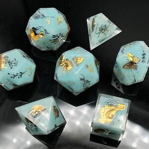 May include: Set of seven teal blue dice with gold accents and various designs. The dice are made of resin and have a translucent finish. The designs include butterflies, flowers, and other nature-inspired motifs.