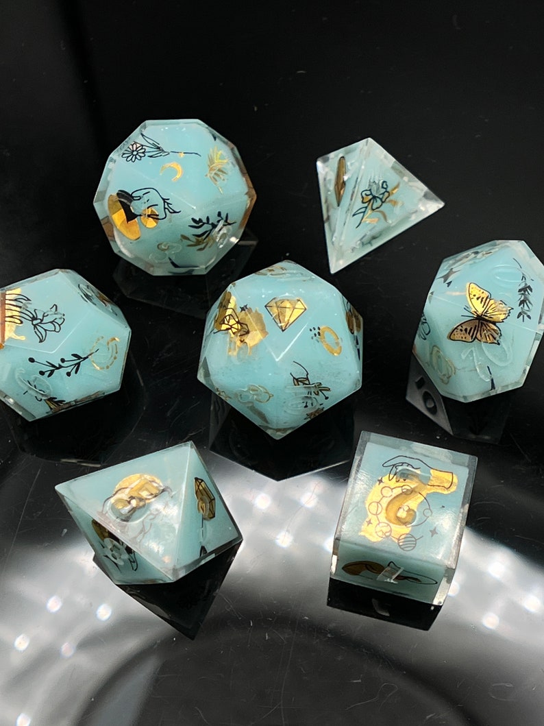May include: Set of seven teal blue dice with gold accents and various designs. The dice are made of resin and have a translucent finish. The designs include butterflies, flowers, and other nature-inspired motifs.