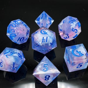 May include: Set of seven iridescent dice with a blue and pink swirl pattern. The dice have a crescent moon and star design with blue numbers.