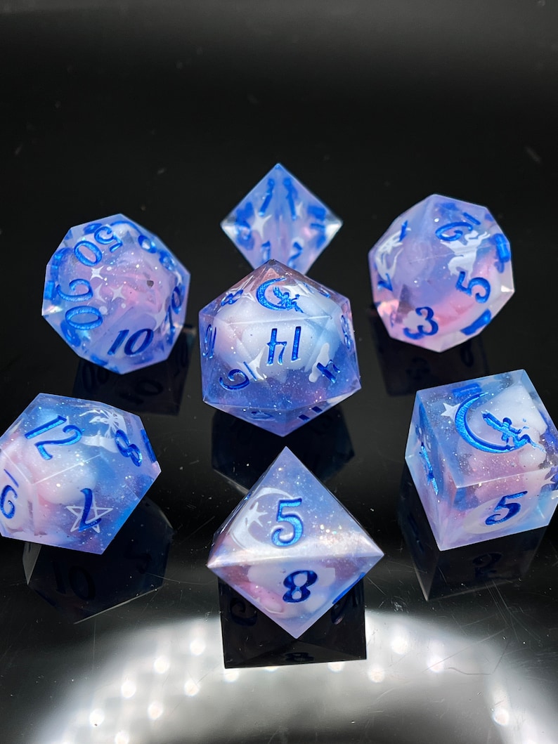 May include: Set of seven iridescent dice with a blue and pink swirl pattern. The dice have a crescent moon and star design with blue numbers.