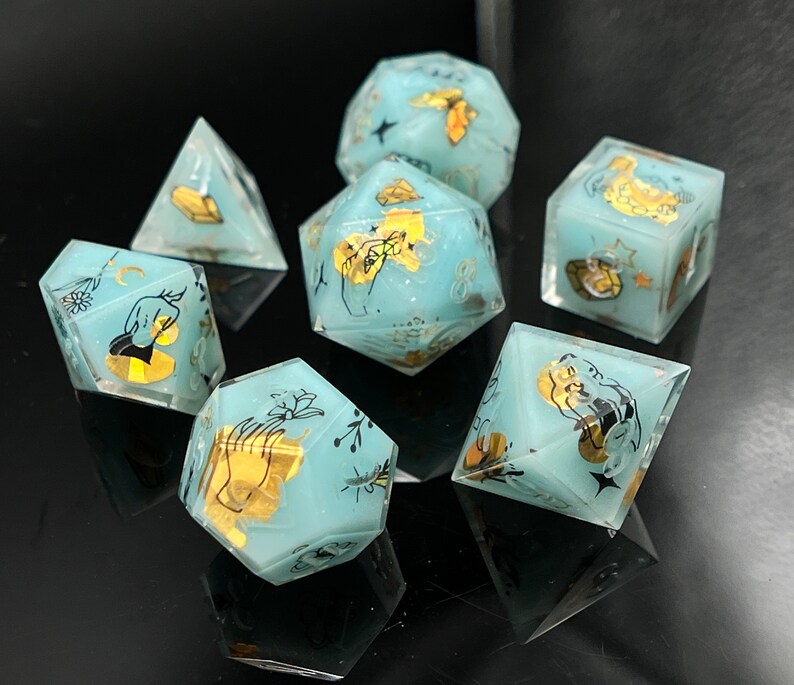 May include: Set of six blue and gold dice with a hand and moon design. The dice are made of resin and have a matte finish.