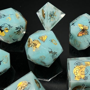May include: Set of eight teal blue dice with gold and black accents. Each die features a different design, including a butterfly, a crescent moon, a hand, and a diamond. The dice are made of a translucent material, allowing the designs to be seen through the dice.