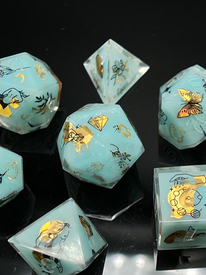 May include: Set of eight teal blue dice with gold and black accents. Each die features a different design, including a butterfly, a crescent moon, a hand, and a diamond. The dice are made of a translucent material, allowing the designs to be seen through the dice.