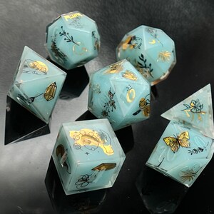 May include: Set of six teal blue dice with gold foil accents and floral designs. The dice are translucent and have a variety of designs, including butterflies, flowers, and crystals.