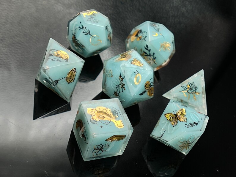 May include: Set of six teal blue dice with gold foil accents and floral designs. The dice are translucent and have a variety of designs, including butterflies, flowers, and crystals.