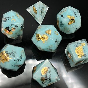 May include: Set of seven teal blue dice with gold accents and various designs, including a butterfly, a crescent moon, a hand, a flower, a diamond, and a bee. The dice are on a black surface.