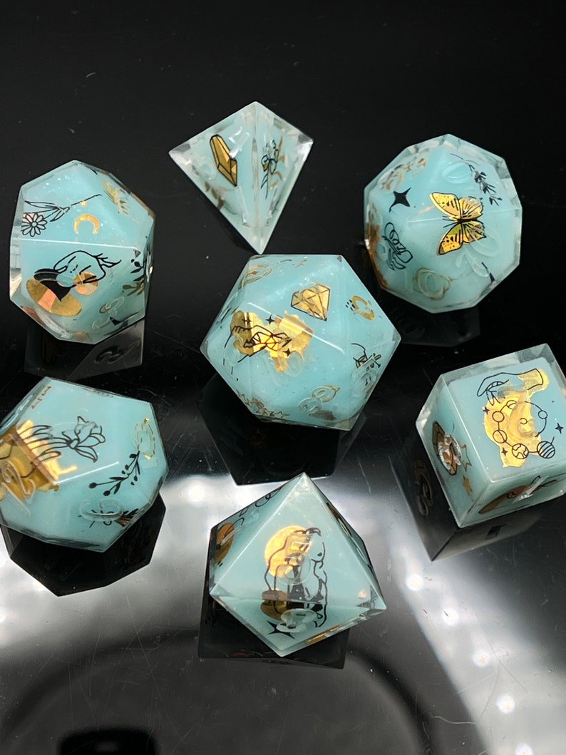 May include: Set of seven teal blue dice with gold accents and various designs, including a butterfly, a crescent moon, a hand, a flower, a diamond, and a bee. The dice are on a black surface.