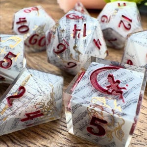 May include: A collection of polyhedral dice with a fantasy theme. The dice are clear with a white interior featuring musical notes and gold accents. The numbers are red. One die has a red crescent moon and fairy design.
