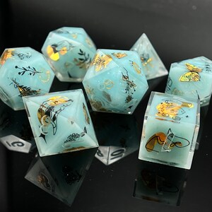May include: Set of seven teal blue dice with gold and black accents. Each die has a different design featuring butterflies, flowers, and other nature-inspired elements.