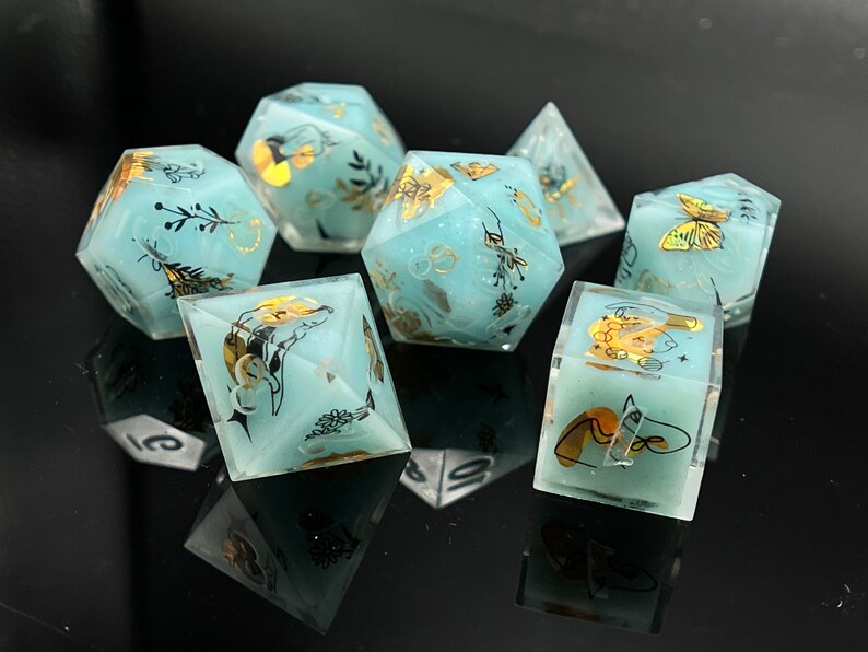 May include: Set of seven teal blue dice with gold and black accents. Each die has a different design featuring butterflies, flowers, and other nature-inspired elements.