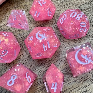 May include: A set of eight pink resin dice with glitter inclusions and light purple numbering. The dice are various shapes, including a d4, d6, d8, d10, d12, d20, and two d10s. Some dice feature a crescent moon and star design.