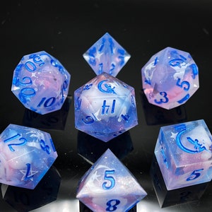 May include: Set of seven iridescent dice with a blue and pink swirl pattern. The dice have white numbers and a crescent moon and star design.