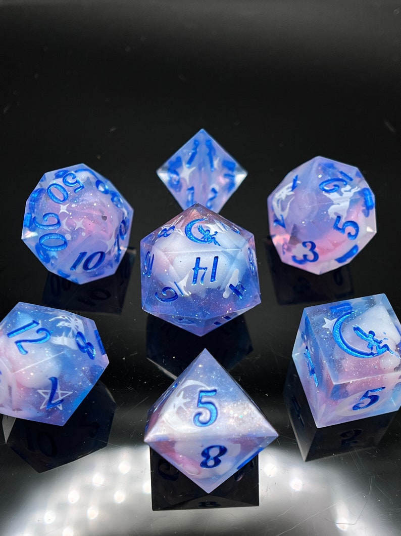 May include: Set of seven iridescent dice with a blue and pink swirl pattern. The dice have white numbers and a crescent moon and star design.