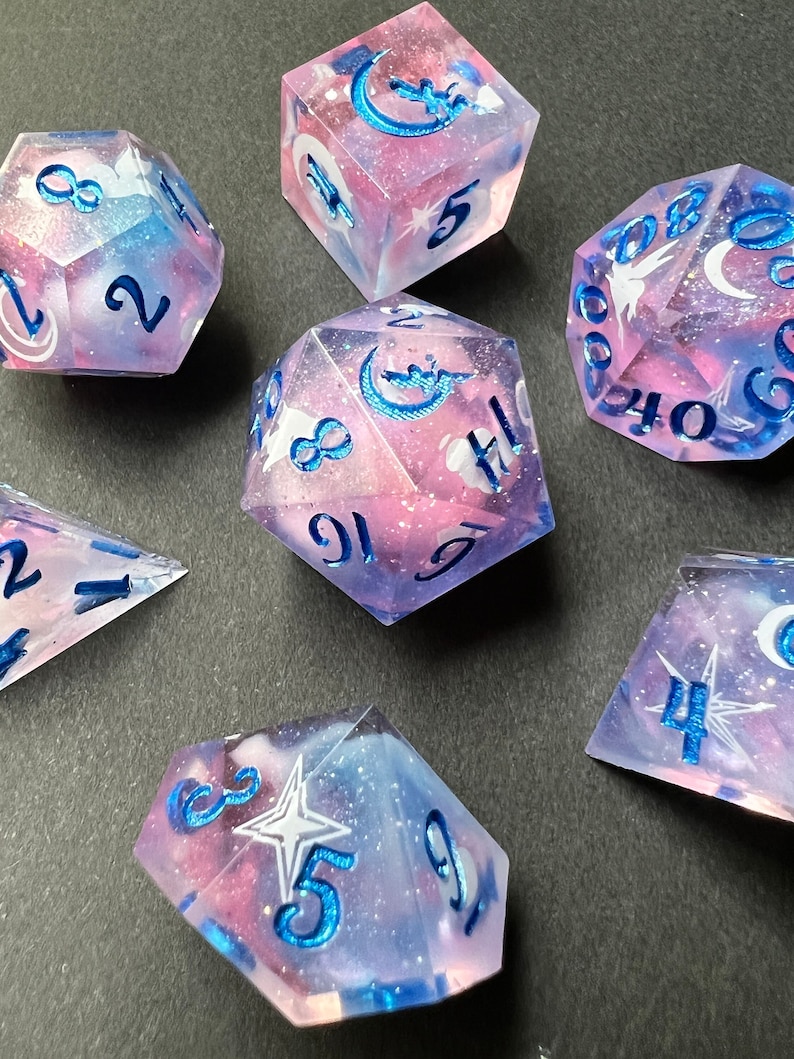May include: Set of seven iridescent dice with a galaxy theme. The dice are pink and blue with glitter and white numbers. The dice have a crescent moon and star design.
