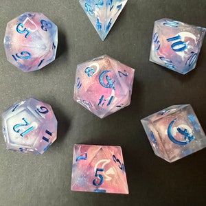 May include: Set of seven iridescent dice with a galaxy-like swirl pattern and blue accents. The dice are numbered 1-6, 1-10, 1-12, 1-20, 1-30, 1-100, and 1-6. The dice are made of clear resin with a galaxy-like swirl pattern and blue accents.