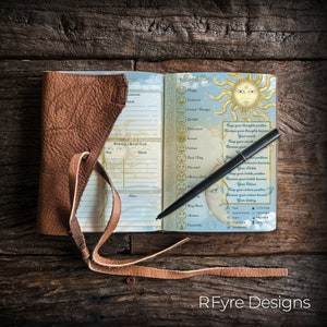 May include: A brown leather-bound journal with a blue and gold interior. The journal has a section for birthdays and special events, a mood tracker, and a section for notes. The journal is open to the mood tracker page, which features a sun with a smiling face and a list of positive affirmations.