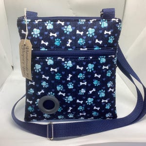 May include: A navy blue crossbody bag featuring a repeating pattern of light blue paw prints, white bones, and small hearts. The bag includes a front zippered pocket, a dark gray circular dispenser opening, and a long adjustable navy strap.
