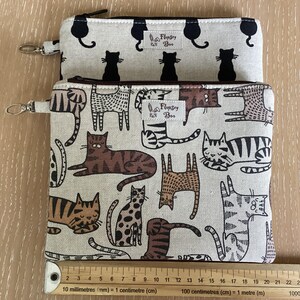 May include: Two rectangular fabric zip pouches with a beige background. The top pouch features black cat silhouettes. The bottom pouch displays various brown and black tabby cat illustrations. Both have a Flopsey Boo label and a metal clip.