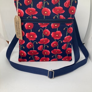 May include: A blue crossbody bag with a red poppy floral print. The bag has two zippered compartments and a navy blue adjustable strap. The tag on the bag reads "Handmade by Hopsey Roo's".