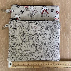 May include: Two beige zipper pouches with black dog illustrations. The top pouch has the word "BOY" printed on it. Both pouches have a silver zipper pull and the "Flopsey Boo" logo. The bottom pouch is covered in dog illustrations.