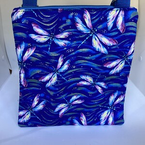 May include: A blue fabric crossbody bag with a zipper closure. The bag features a repeating pattern of colorful dragonflies on a blue background with gold accents.