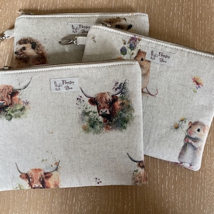 May include: Three zipped pouches with animal illustrations. The pouches are made from a light beige linen-like fabric. One pouch features a hedgehog, another a Highland cow, and the third a mouse. Each pouch has a silver zip and a label that says "Flopsy & Bee".