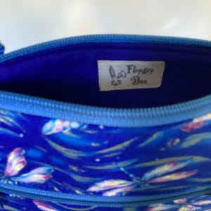 May include: The inside of a blue zippered pouch with a blue fabric lining. The pouch has a white label with the text "Flopsy Bee" in brown lettering.
