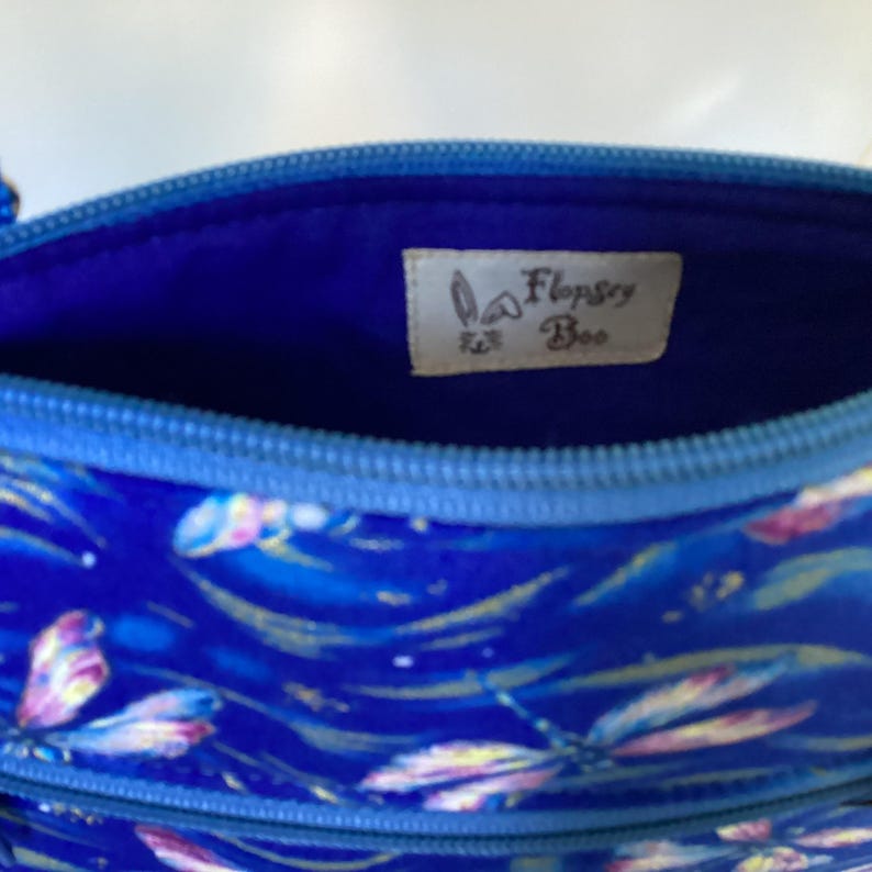May include: The inside of a blue zippered pouch with a blue fabric lining. The pouch has a white label with the text "Flopsy Bee" in brown lettering.