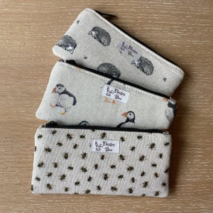 May include: Three canvas zipper pouches with nature-themed designs. One pouch features hedgehogs, another puffins, and the third has a pattern of bees. Each pouch has a black zipper and a small label that reads "Flopsy Boo".