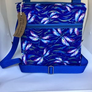 May include: A blue crossbody bag with a dragonfly print and a blue strap. The bag has two zippered compartments and a tag that reads "Handmade by Flopsy Boo".