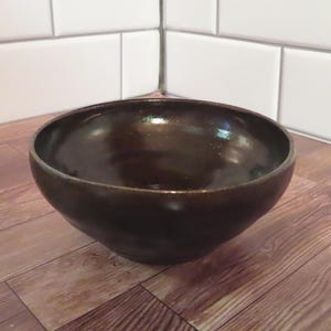May include: A deep, round ceramic bowl with a smooth, dark brown glazed finish. The bowl features a wide, open top and a slightly tapered base, showcasing a reflective, glossy surface that catches the light across its curved interior and exterior.