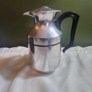 May include: A silver and black coffee pot with a lid and a handle. The pot is sitting on a white fabric surface.