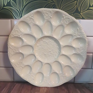 Op de afbeelding: Een ronde, crèmekleurige keramische eierserveerschaal met een centraal rond gedeelte omringd door achttien ovale uitsparingen. Het oppervlak is versierd met een verhoogd, gestructureerd botanisch patroon van bladeren en druiven.