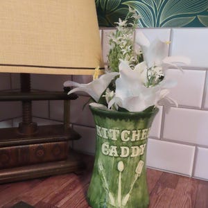 May include: A green ceramic kitchen caddy with a flared rim and base. The front features the embossed text KITCHEN CADDY above raised illustrations of a fork, spoon, and spatula. The caddy is filled with a bouquet of white artificial flowers.