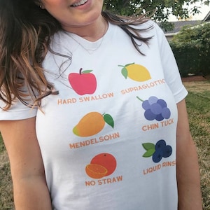 Può includere: Maglietta bianca con un design di frutta colorata e testo. Il testo include "Hard Swallow", "Supraglottic", "Mendelsohn", "Chin Tuck", "No Straw" e "Liquid Rinse".