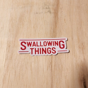 Puede incluir: Pegatina blanca con texto rojo que dice "SWALLOWING THINGS" con una línea arriba y abajo del texto.