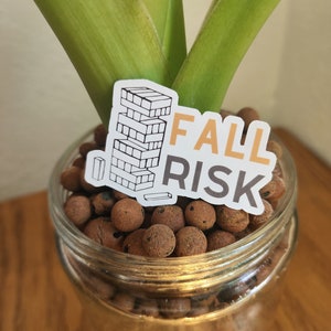 Puede incluir: Una pegatina blanca con el texto "FALL RISK" y una ilustración en blanco y negro de una torre de bloques. La pegatina está colocada en un frasco de vidrio lleno de bolitas de arcilla marrones.