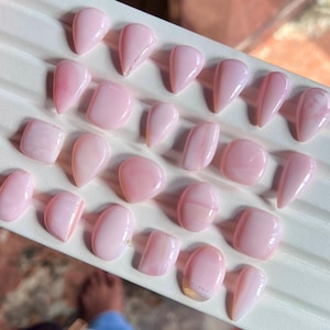May include: A selection of pink opal gemstones in diverse forms, such as teardrops, ovals, and rectangles. The polished stones exhibit a soft, translucent quality with delicate colour variations and internal patterns. The gemstones are displayed on a white surface.