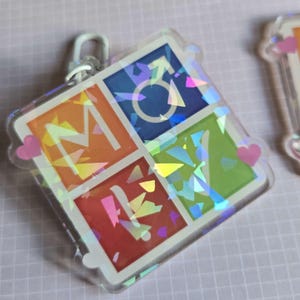 Pode incluir: Um chaveiro quadrado com borda transparente e fecho prateado. O chaveiro &eacute; dividido em quatro quadrados coloridos, cada um com uma letra que forma "MOVE" e um s&iacute;mbolo. Os quadrados s&atilde;o laranja, azul, vermelho e verde, com um efeito hologr&aacute;fico.