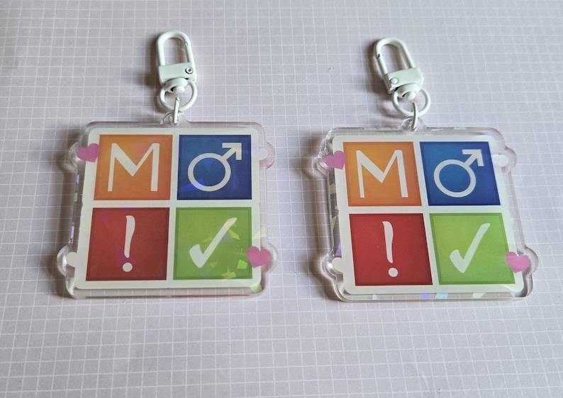 Pode incluir: Dois chaveiros quadrados com fecho de metal branco. Cada chaveiro tem um design colorido com as letras "MO", um s&iacute;mbolo masculino, um ponto de exclama&ccedil;&atilde;o e uma marca de verifica&ccedil;&atilde;o. Acentos de cora&ccedil;&atilde;o rosa nos cantos.