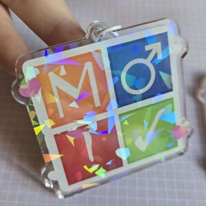 Pode incluir: Um chaveiro de acr&iacute;lico transparente com efeito hologr&aacute;fico. O chaveiro apresenta quatro quadrados coloridos: laranja com a letra "M", azul com um s&iacute;mbolo masculino, vermelho com a letra "T" e verde com uma marca de verifica&ccedil;&atilde;o.