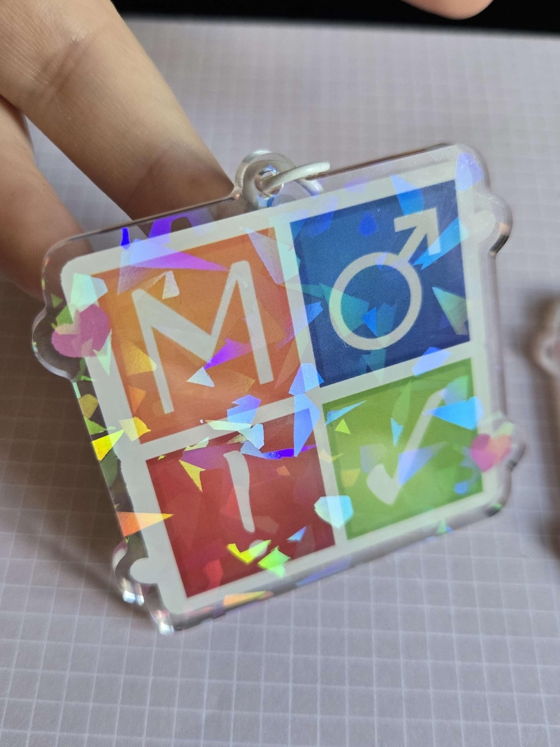 Pode incluir: Um chaveiro de acr&iacute;lico transparente com efeito hologr&aacute;fico. O chaveiro apresenta quatro quadrados coloridos: laranja com a letra "M", azul com um s&iacute;mbolo masculino, vermelho com a letra "T" e verde com uma marca de verifica&ccedil;&atilde;o.