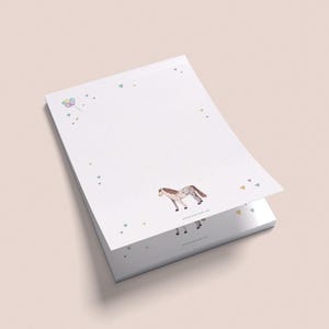 Könnte beinhalten: Ein Stapel weißes Notizblockpapiers mit einem Pferde- und Schmetterlingsdesign. Das Papier hat einen hellrosa Hintergrund mit kleinen bunten Punkten.