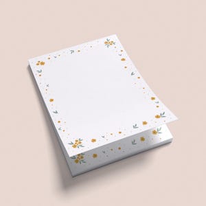 Könnte beinhalten: Ein Stapel weißer Briefpapierbögen mit einem floralen Rand. Der Rand zeigt kleine gelbe Blumen und grüne Blätter. Das Briefpapier liegt auf einer hellrosa Oberfläche.