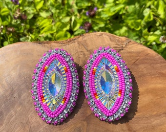 Puede incluir: Un par de pendientes de cuentas rosas y blancas con un cristal transparente iridiscente en el centro. Los pendientes son de forma ovalada y tienen un respaldo plateado.