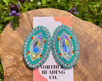 Puede incluir: Pendientes ovalados de cuentas turquesa y plata con un centro de cristal. Los pendientes tienen un borde de cuentas rojas y amarillas. Los pendientes se muestran sobre una superficie de madera con una tarjeta que dice "Norther Beading Co."