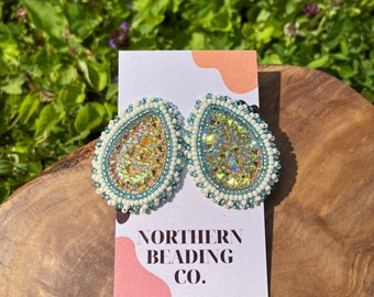 May include: A pair of iridescent teardrop-shaped earrings with white and turquoise beads. The earrings are on a white card with the text "NORTHERN BEADING CO."