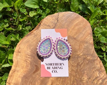 May include: A pair of iridescent teardrop-shaped earrings with purple bead accents. The earrings are on a wooden surface with a business card that reads "Northern Beading Co."